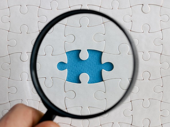 Hand holds magnifying glass over a jigsaw puzzle with a piece missing